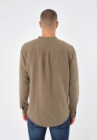 Pitkähihainen paita oliivinvihreästä kankaasta, jossa on selkäyoke, pyöristetty helma ja napilliset kalvosimet, yhdistettynä sinisiin farkkuihin.