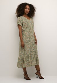 Cvečni midi obleka v zeleni barvi s rožami v rožnati in rumeni barvi, kratkimi nabrano rokavi, elastičnim pasom in večplastnim robom. Utemeljena s črnimi sandali na zavezo.