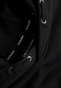 Close-up af sort AllSaints hættetrøje, der viser snøreløg og indvendigt mærke med AllSaints branding på stofbånd.