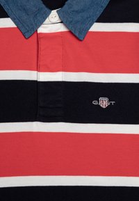 Rød, hvit og marineblå stripete polo skjorte med jeanskrage. Har en tre-knapp placket og en liten brodert logo på brystet.