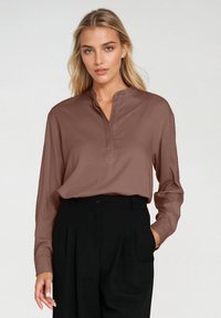 Brązowa koszula z długim rękawem, z kołnierzykiem stojącym i trzema guzikami, stylizowana z czarnymi spodniami o wysokim stanie. Gładka faktura materiału.