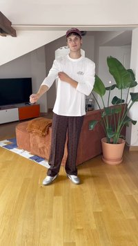 Giovane uomo che indossa una camicia bianca a maniche lunghe, pantaloni a fantasia, scarpe argentate e un cappello bordeaux, in piedi in un soggiorno con piante e un televisore.