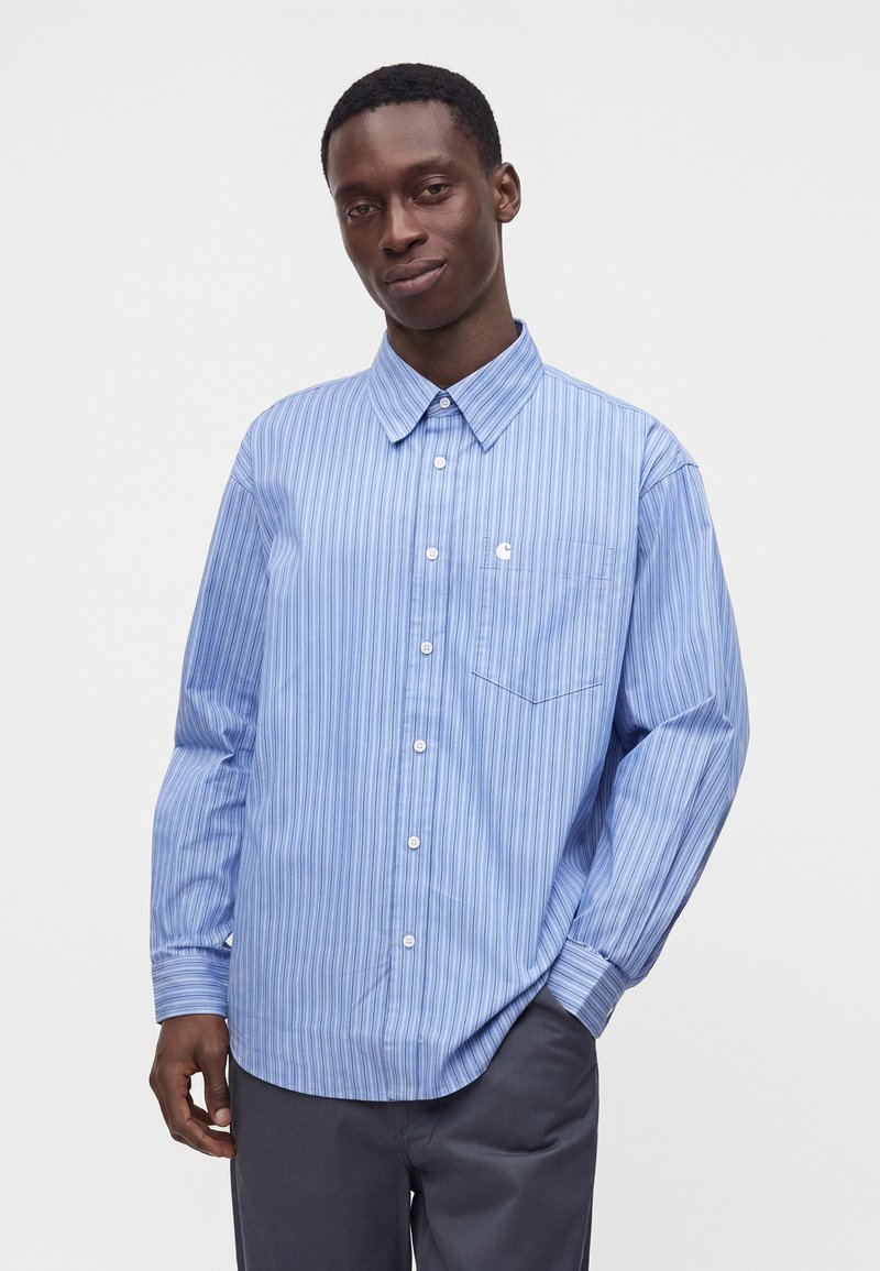 Homme portant une chemise boutonnée bleu clair à rayures verticales et un pantalon sombre, debout devant un fond clair uni.