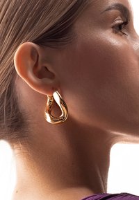 Orecchini in metallo color oro con una forma fluida e astratta a goccia, che riflettono la luce con una finitura lucida, indossati all'orecchio di una persona.