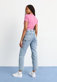 Rosa kortermet t-skjorte, høyt liv lyseblå jeans med lommer, og hvite sneakers på en person som står på en blå overflate.