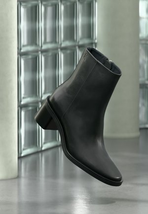 Black leather ankle boot with low block heel and zipper, set against a glass block wall and concrete floor background.