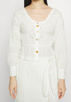 Kobieta ubrana w białą, fakturowaną bluzkę z długimi rękawami, dekoltem w serek, złotymi i białymi guzikami oraz dopasowaną spódnicę z wiązanym paskiem w talii.