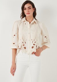 Blusa color crema con bordado floral recortado, frente con botones y manga tres cuartos. El material parece ligero y texturizado.