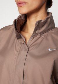 Smeđa lagana jakna s visokim ovratnikom, sadrži srebrni Nike logotip na lijevoj strani. Glatka tekstura i detalj s patentnim zatvaračem.
