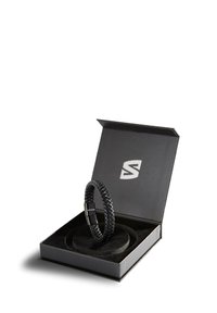 Black braided leather bracelet with metallic clasp inside black open box with a padded interior and a white logo on the lid.