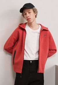 Felpa rossa con cerniera e dettagli neri, t-shirt bianca sotto e pantaloni scuri. Presenta un cappuccio e tasche laterali con zip.