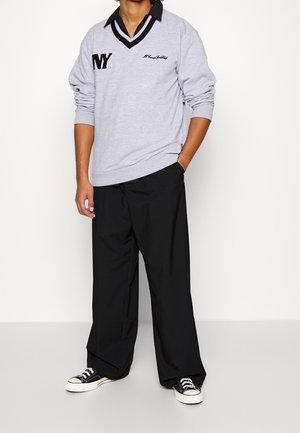 Persona in maglione lungo grigio con colletto nero e testo "NY", pantaloni neri larghi e sneakers in bianco e nero, in piedi contro una parete bianca.