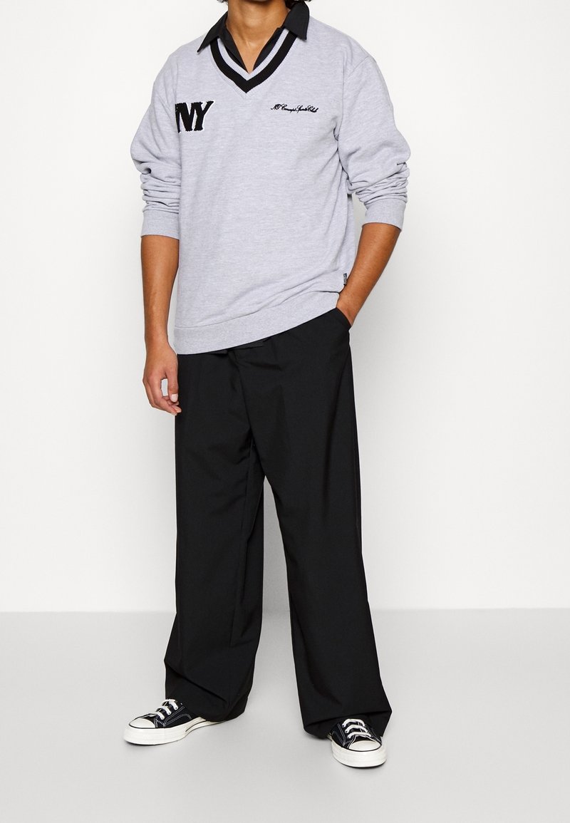 Persona in maglione lungo grigio con colletto nero e testo "NY", pantaloni neri larghi e sneakers in bianco e nero, in piedi contro una parete bianca.
