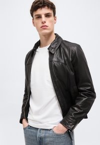 Νεαρός άνδρας με τα χέρια στις τσέπες, φοράει μαύρο δερμάτινο μπουφάν πάνω από λευκό πουκάμισο και μπλε τζιν, μπροστά σε απλό φωτεινό φόντο.