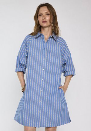 Femme debout, les mains dans les poches, portant une robe chemise à rayures verticales bleues et blanches avec les manches retroussées, sur un fond uni.