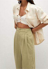 Chemise beige cintrée à boutons portée sur un débardeur blanche à côtes avec des boutons ; pantalon vert clair taille haute et plissé avec poches latérales.