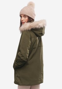 Parka verde oliva con cappuccio bordato di pelliccia, maniche lunghe, tasche frontali e superficie liscia. Berretto beige a maglia con pom-pom sulla sommità.