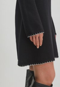 Schwarzes Strickkleid mit langen Ärmeln, das eine strukturierte Webart aufweist. Auffällige Details sind der kontrastierende weiße gewellte Saum und die Ärmelabschlüsse.