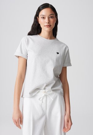 Woman with long dark hair wearing a light gray short-sleeve t-shirt with a small black heart emblem and white drawstring pants.