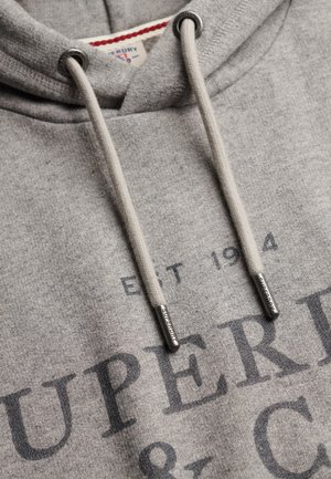 Zbliżenie na szarą bluzę z kapturem ze sznurkami, na których znajdują się metalowe zakończenia z wygrawerowanym napisem "Superdry" oraz częściowo widoczny nadruk z tekstem "EST 1984 SUPERD".