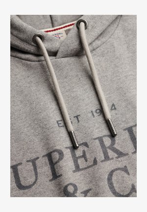 Zbliżenie na szarą bluzę z kapturem ze sznurkami, na których znajdują się metalowe zakończenia z wygrawerowanym napisem "Superdry" oraz częściowo widoczny nadruk z tekstem "EST 1984 SUPERD".