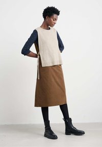 Beige kötött mellény egy sötétkék, pöttyös, hosszú ujjú felsőn, barna midi szoknyával és fekete fűzős csizmával párosítva. A szoknyán nincsenek látható minták.