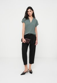 Πράσινο τοπ με κοντά μανίκια και V-λαιμόκοψη, συνδυασμένο με μαύρο παντελόνι με στενή γραμμή. Το μοντέλο κρατά μικρό μπορντό τσαντάκι με ασημένιες λεπτομέρειες. Μαύρα παπούτσια με λουράκι στο πίσω μέρος.