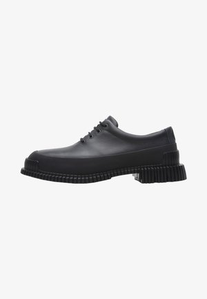 Zapato de goma negro con punta redondeada, parte superior suave y suela gruesa estriada. Cuenta con cinco ojales para los cordones y un diseño robusto.