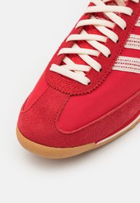 Röd sneaker med en kombination av slät tyg och mocka, rund tå, vita snören och en sula av gummi med subtil textur.