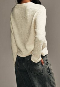 Pull en maille texturée couleur crème avec une coupe décontractée, porté avec un jean gris foncé taille haute, doté de poches arrière et d'un léger effet usé.