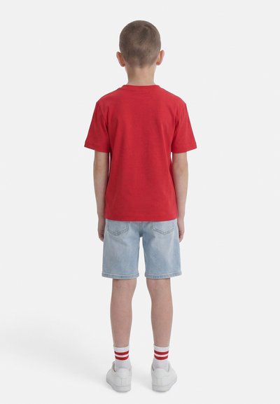 Αγόρι που στέκεται με την πλάτη γυρισμένη, φοράει κόκκινο μπλουζάκι, ανοιχτό μπλε τζιν σορτς, λευκά αθλητικά παπούτσια και λευκές κάλτσες με κόκκινες ρίγες.