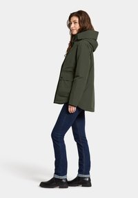 Olijfgroene hoodie-jas met een rits aan de voorkant, twee zijzakken en een relaxte pasvorm. G gedragen met donkere spijkerbroek en zwarte enkellaarsjes.