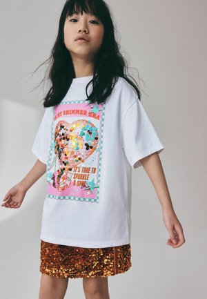 Mädchen trägt weißes oversized T-Shirt mit buntem Herzmotiv und bronzenem Paillettenrock und steht vor einfarbigem Hintergrund.