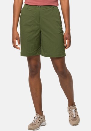 Persona que lleva puestos pantalones cortos de color verde oliva hasta la rodilla con bolsillos laterales y zapatos deportivos beige, de pie frente a un fondo liso.
