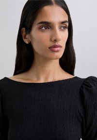 Woman with long dark hair wearing a textured black top with puffed sleeves, looking slightly right against a plain light background.