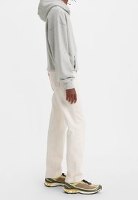 Sudadera con capucha gris, pantalones rectos beige y zapatillas deportivas verde/dorado. El conjunto tiene un ajuste relajado y casual, con tonos neutros.