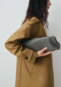 Grå läderclutch som hålls under armen, med en rundad form och subtila sömdetaljer, i kombination med en brun kappa med stor krage.