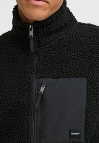 Black fleece jacket featuring a high collar, zippered front, and a contrasting black pocket with a logo label. Soft, textured fabric.