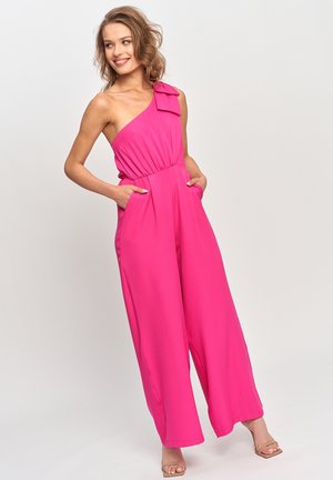 Kobieta uśmiechająca się, ubrana w jasnoróżowy kombinezon na jedno ramię z szerokimi nogawkami, z rękami w kieszeniach i beżowe sandały na obcasie.