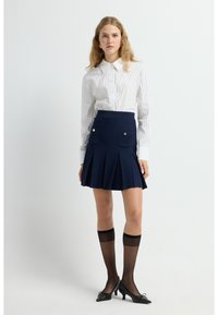 Camisa de botones blanca a rayas, falda plisada azul marino con bolsillos y detalles dorados, combinada con medias altas negras transparentes y tacones negros.