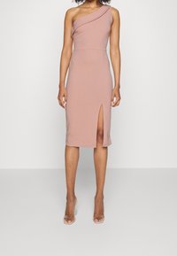 Femme portant une robe midi mauve sans manches et à épaule unique avec une fente à l'avant, associée à des sandales à talons transparents.