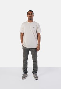 Beige T-Shirt mit kleinem schwarzen Elefanten-Logo, kombiniert mit grünen Hosen und grauen Sneakers. Model in einer neutralen Umgebung präsentiert.