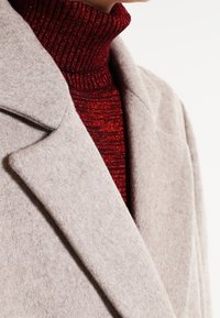 Lichtgrijze wollen jas met notch-lapels, gelaagd over een getailleerde, geribbelde coltrui in rood met metallic accenten. Close-up van texturen en gelaagdheid.