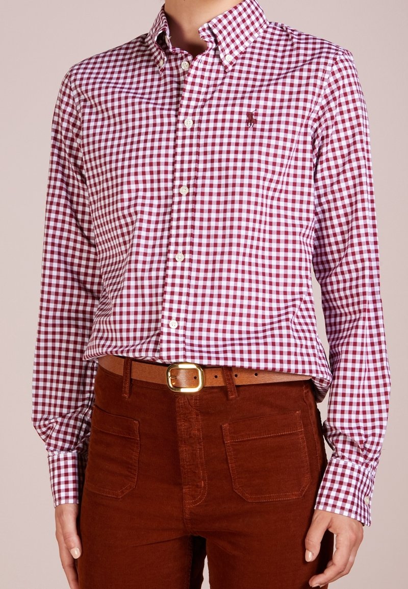 Camicia a scacchi rossa e bianca con maniche lunghe, dotata di colletto e un piccolo emblema sul petto, abbinata a pantaloni di velluto a coste marroni.