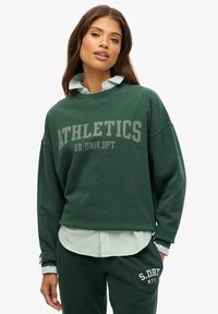 Sudadera verde con el texto "ATHLETICS", corte relajado, cuello redondo, superpuesta sobre una camiseta de rayas azul claro, combinada con pantalones verdes a juego.