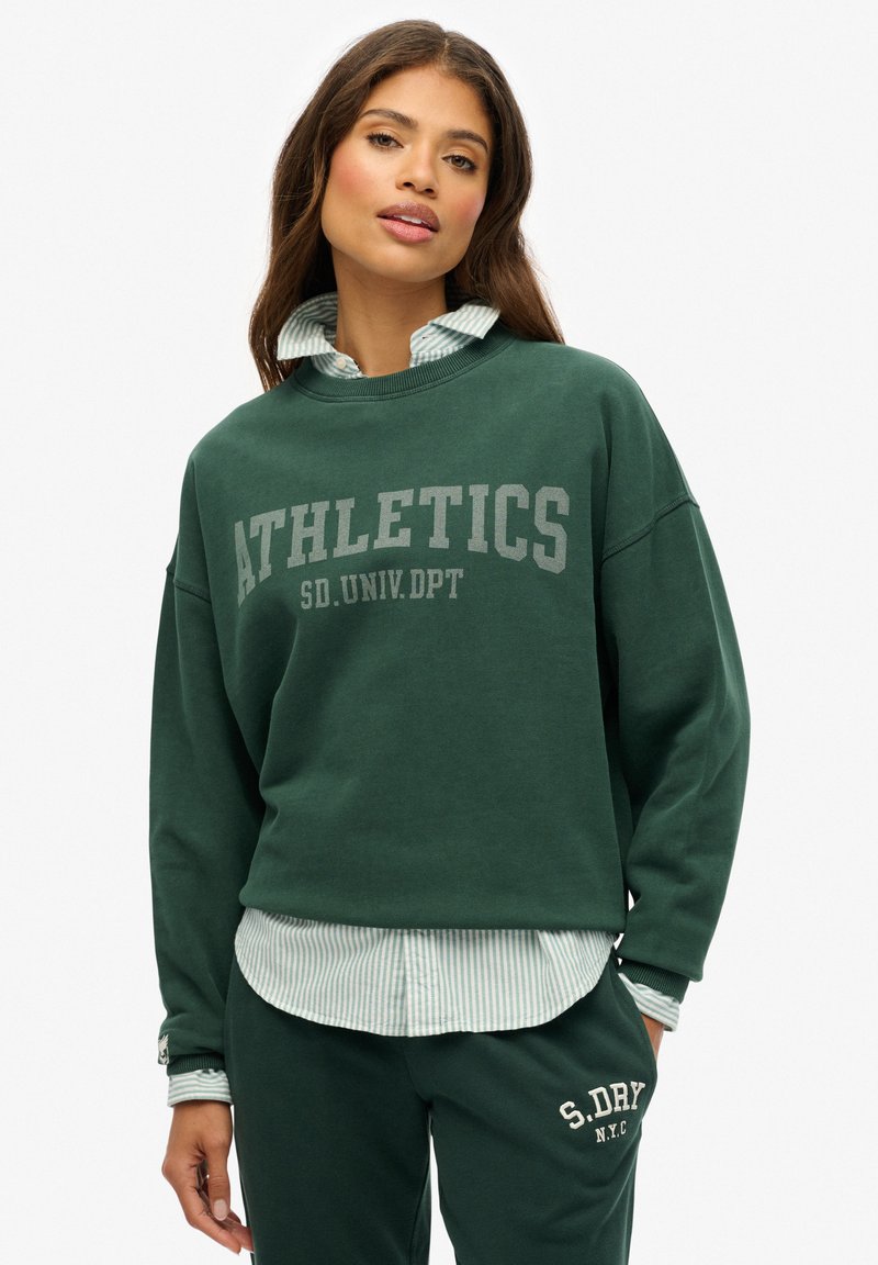Sudadera verde con el texto "ATHLETICS", corte relajado, cuello redondo, superpuesta sobre una camiseta de rayas azul claro, combinada con pantalones verdes a juego.