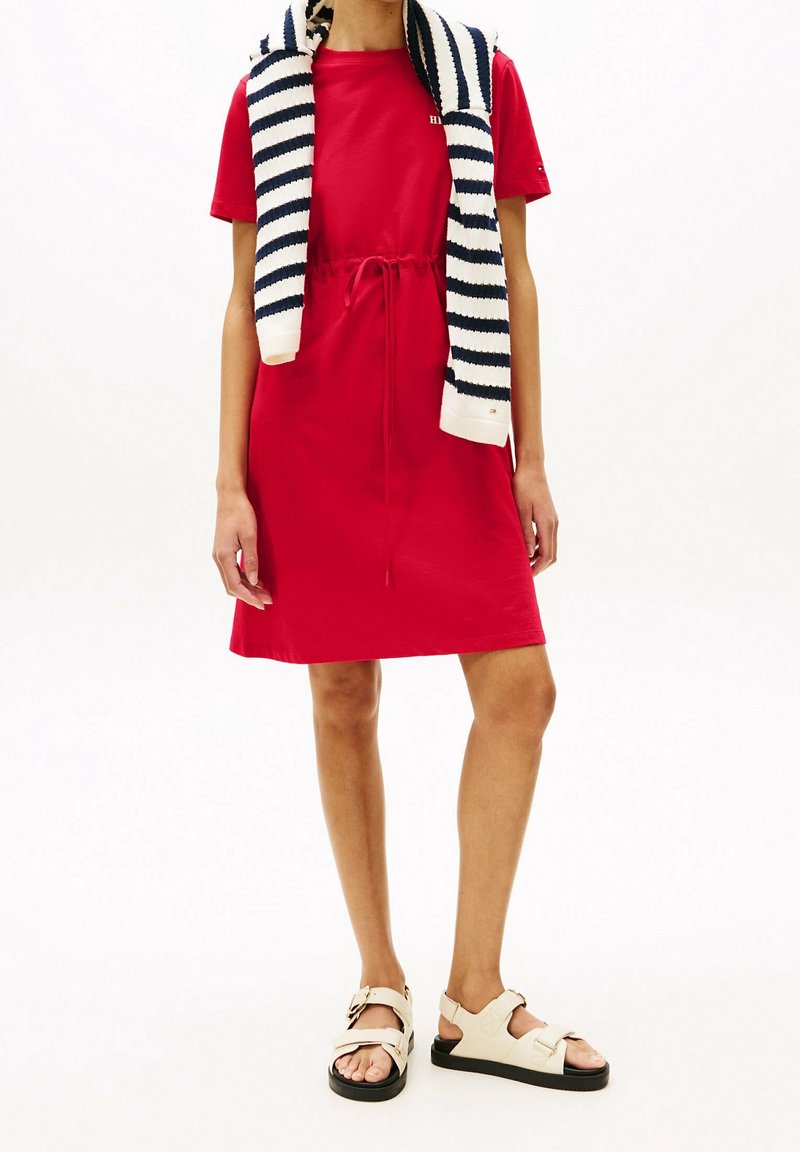 Vestido de algodón rojo con cintura ajustable. Combinado con un suéter de rayas en azul marino y blanco que cuelga sobre los hombros. Lleva sandalias beige.