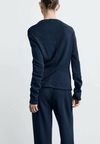 Maglia a maglia blu scuro con scollo rotondo e maniche lunghe, caratterizzata da un dettaglio di cucitura sul retro. Abbinata a pantaloni scuri coordinati.