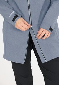 Light blue jacket with a zip front, featuring a textured fabric, black accents, and a thumbhole design at the cuffs. Worn over black pants.