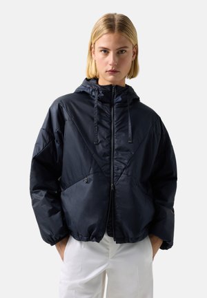 Jasnowłosa osoba ubrana w czarną pikowaną kurtkę z kapturem, z przodu zamykaną na zamek i z kieszeniami, w połączeniu z białymi spodniami, stojąca na jednolitym tle.